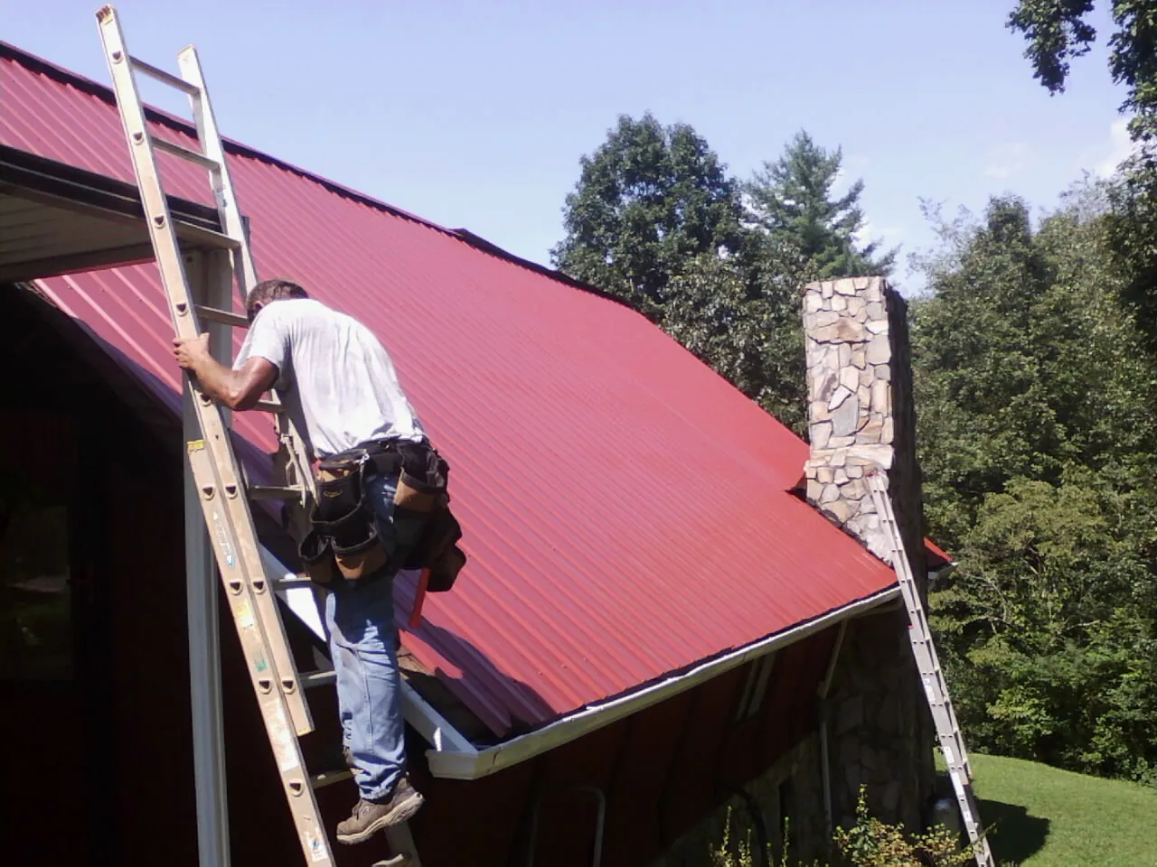 Roofer on a ladder beside a red metal roof and stone chimney — Soffit & Fascia Repair by Roofing Brothers in Bound Brook